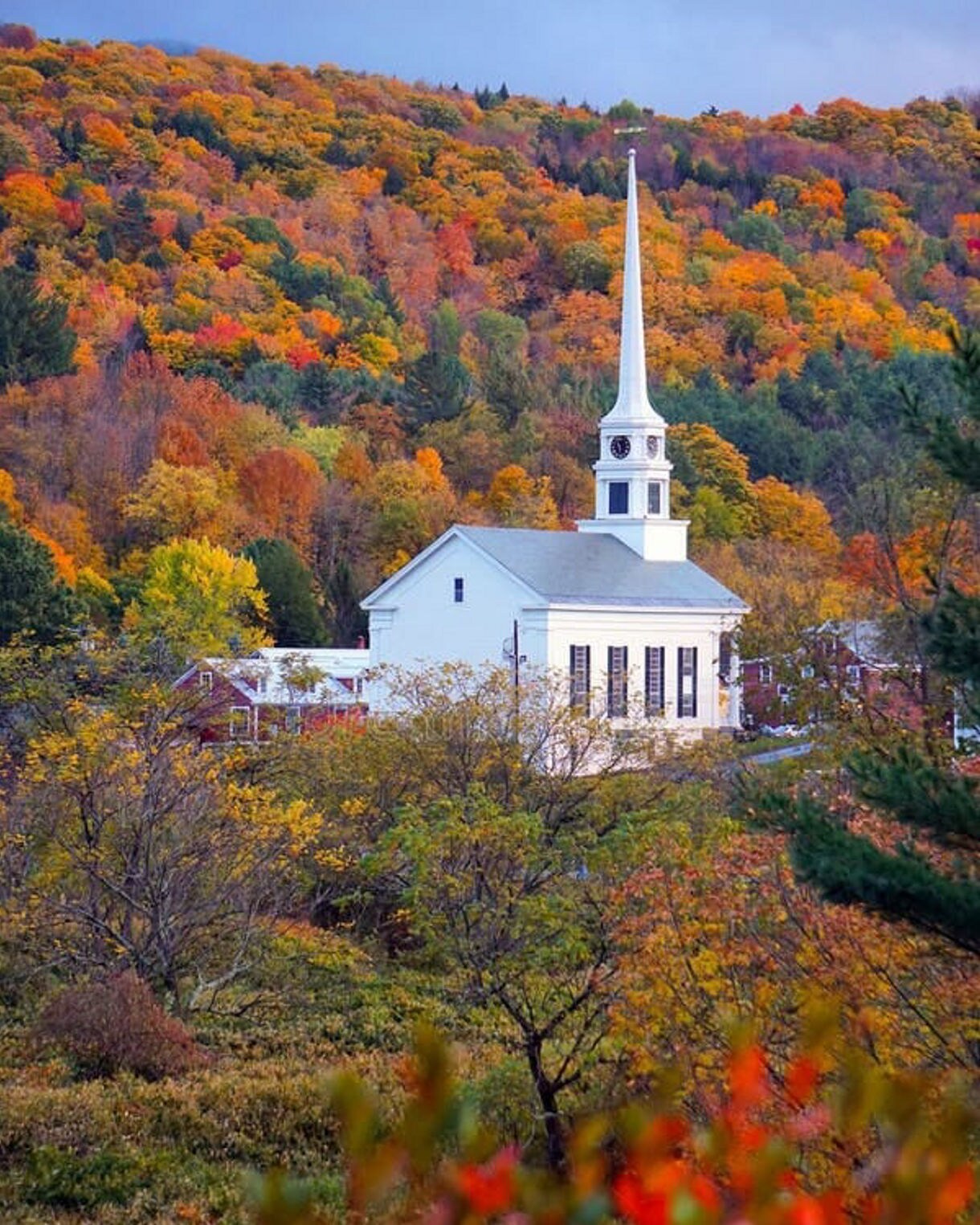 Classic white church with a tall steeple surrounded by vibrant autumn foliage covering a hillside under soft evening light.