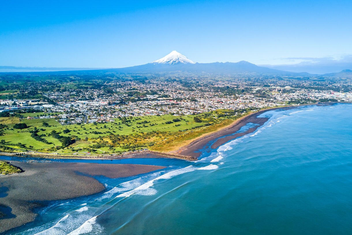 Aerial view of New Plymouth’s coastline with rolling waves, green fields and Mount Taranaki’s snowy peak in the distance beneath a clear blue sky.