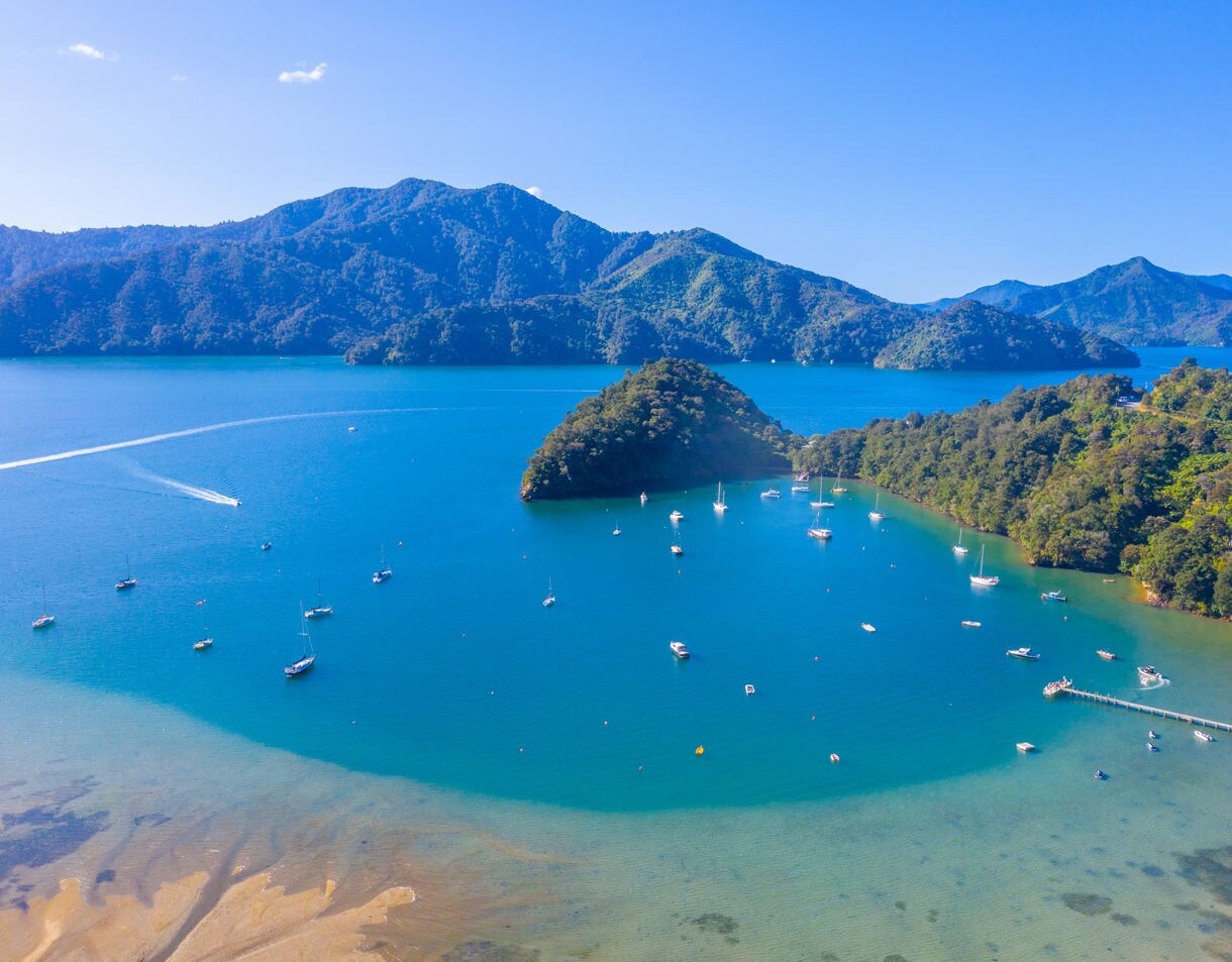 Aerial view of Ngakuta Bay with bright blue water, anchored sailboats and forested hills rising in the background.