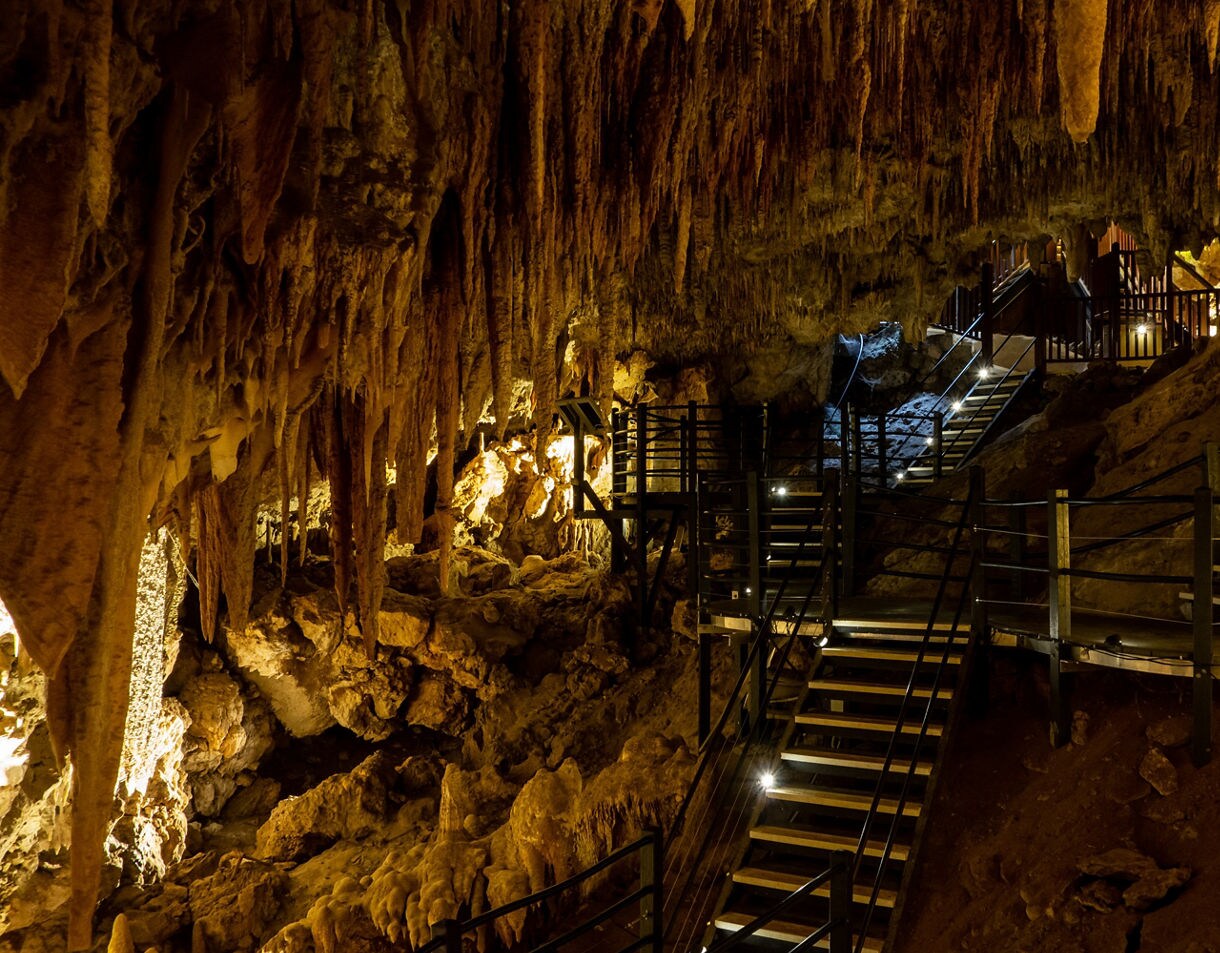  Interior of a limestone cave with dramatic stalactites hanging from the ceiling and a series of lit walkways and stairs leading through the cavern.