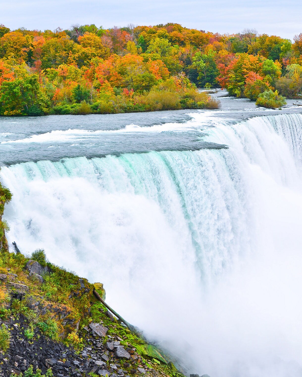 Aerial view of Niagara Falls surrounded by orange and green autumn trees, with mist rising from the powerful rushing waterfall below.