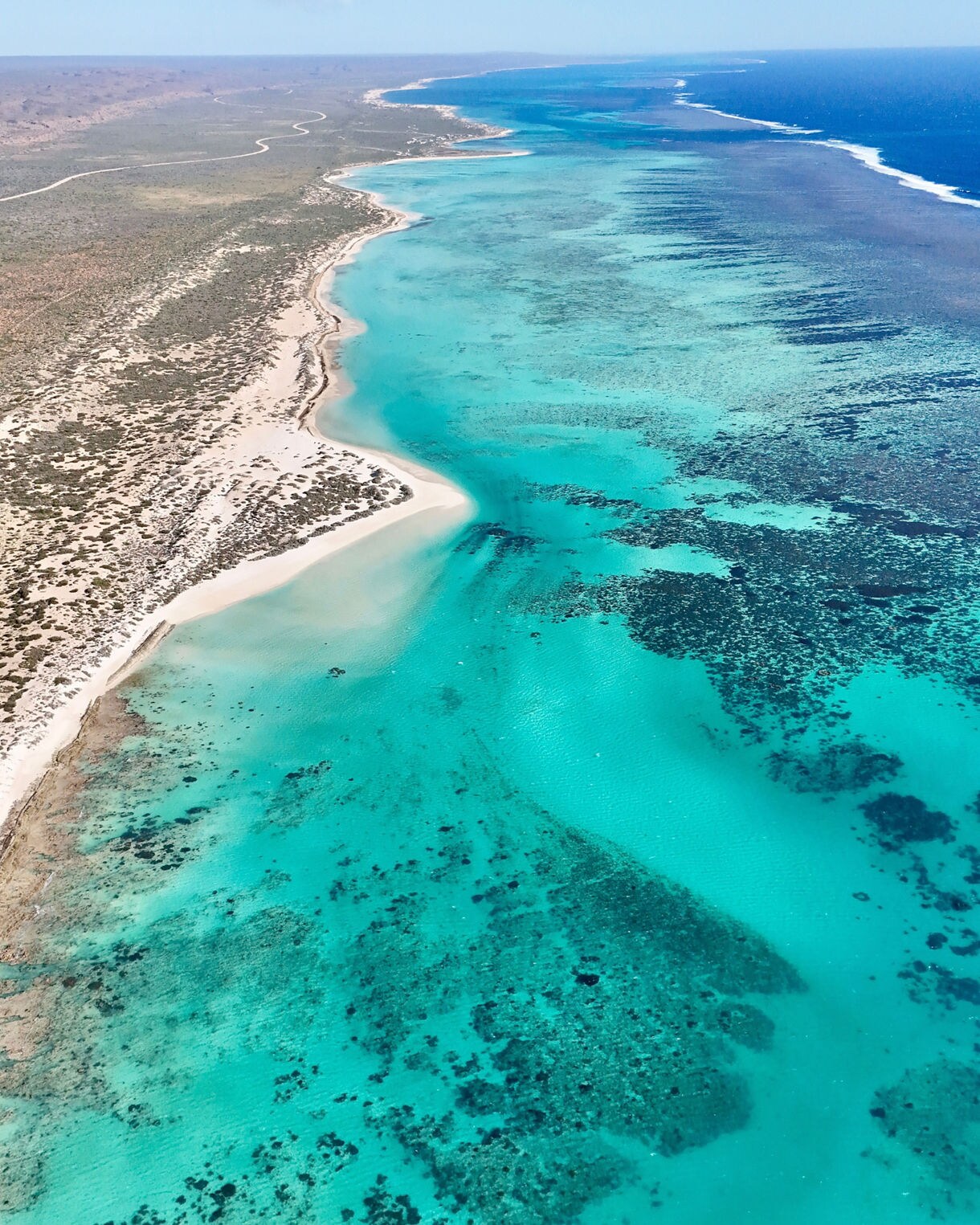 Aerial view of the Ningaloo Reef coastline showing bright turquoise water, coral patches and a long curve of white sand backed by scrubby desert.