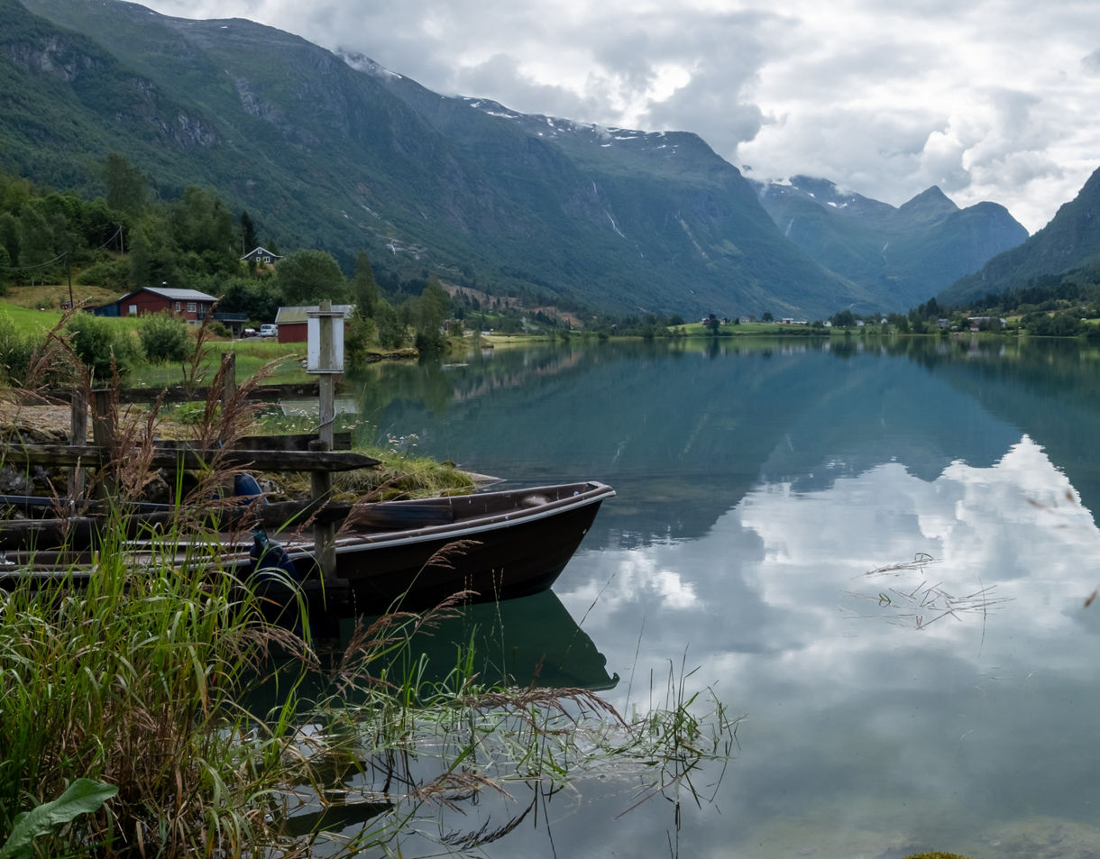 Nordfjord in Norway with glassy water reflecting mountains, scattered farmhouses and moored wooden boats in the foreground.