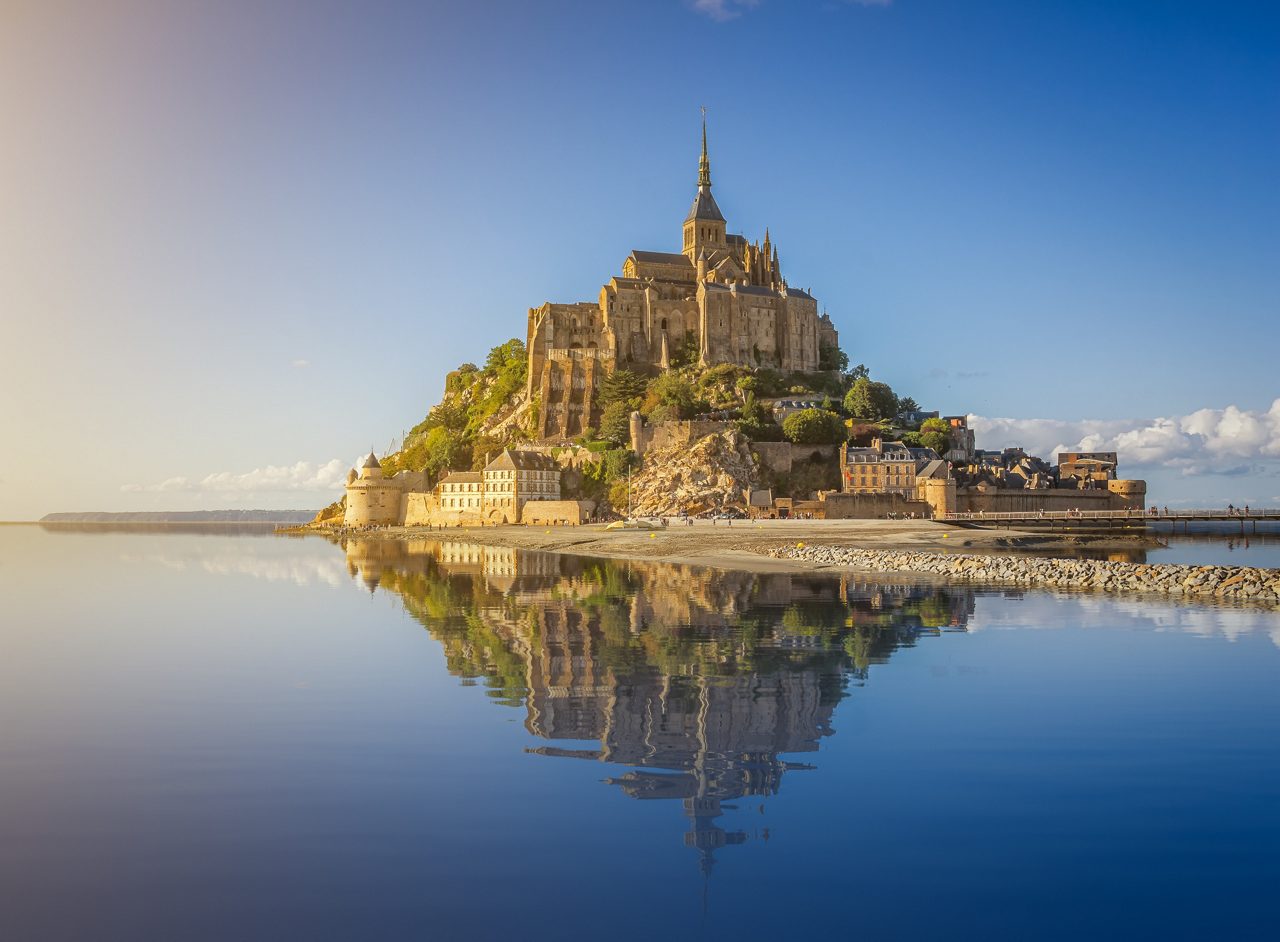 A stunning view of Mont Saint-Michel in Normandy, France, with the ancient abbey perched on a rocky island, reflected in the calm water under a clear blue sky.