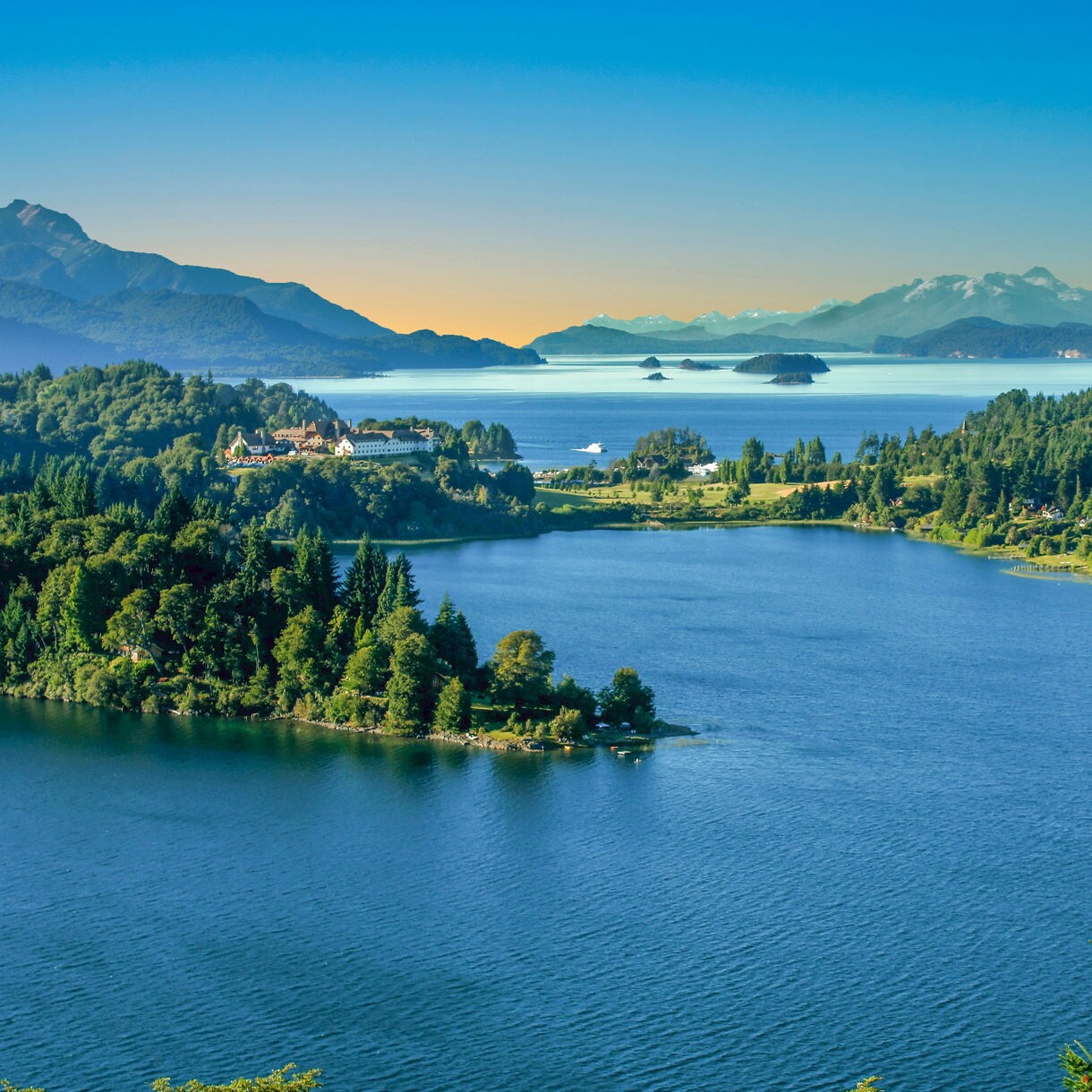 Elevated view of a large blue lake surrounded by forested hills with distant mountains and scattered small islands under a clear sky.