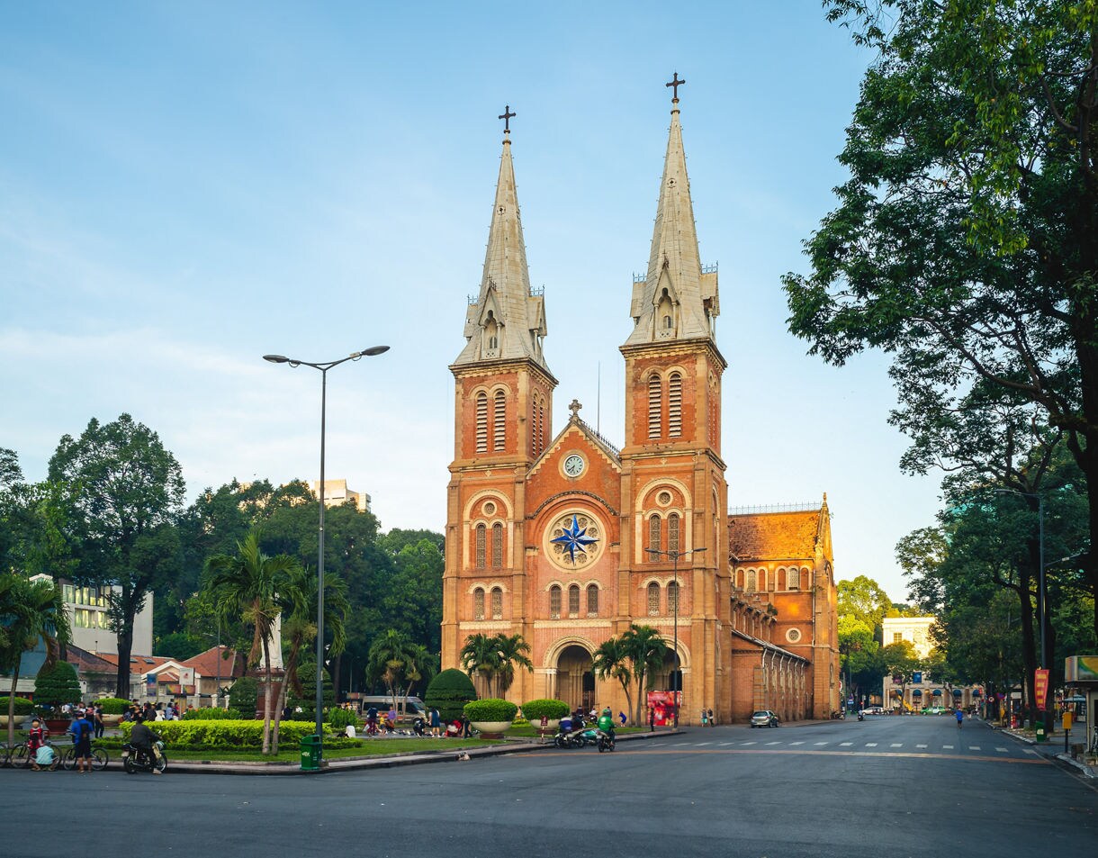Red-brick Notre-Dame Cathedral Basilica of Saigon with twin bell towers and a blue rose window, surrounded by trees and city streets.