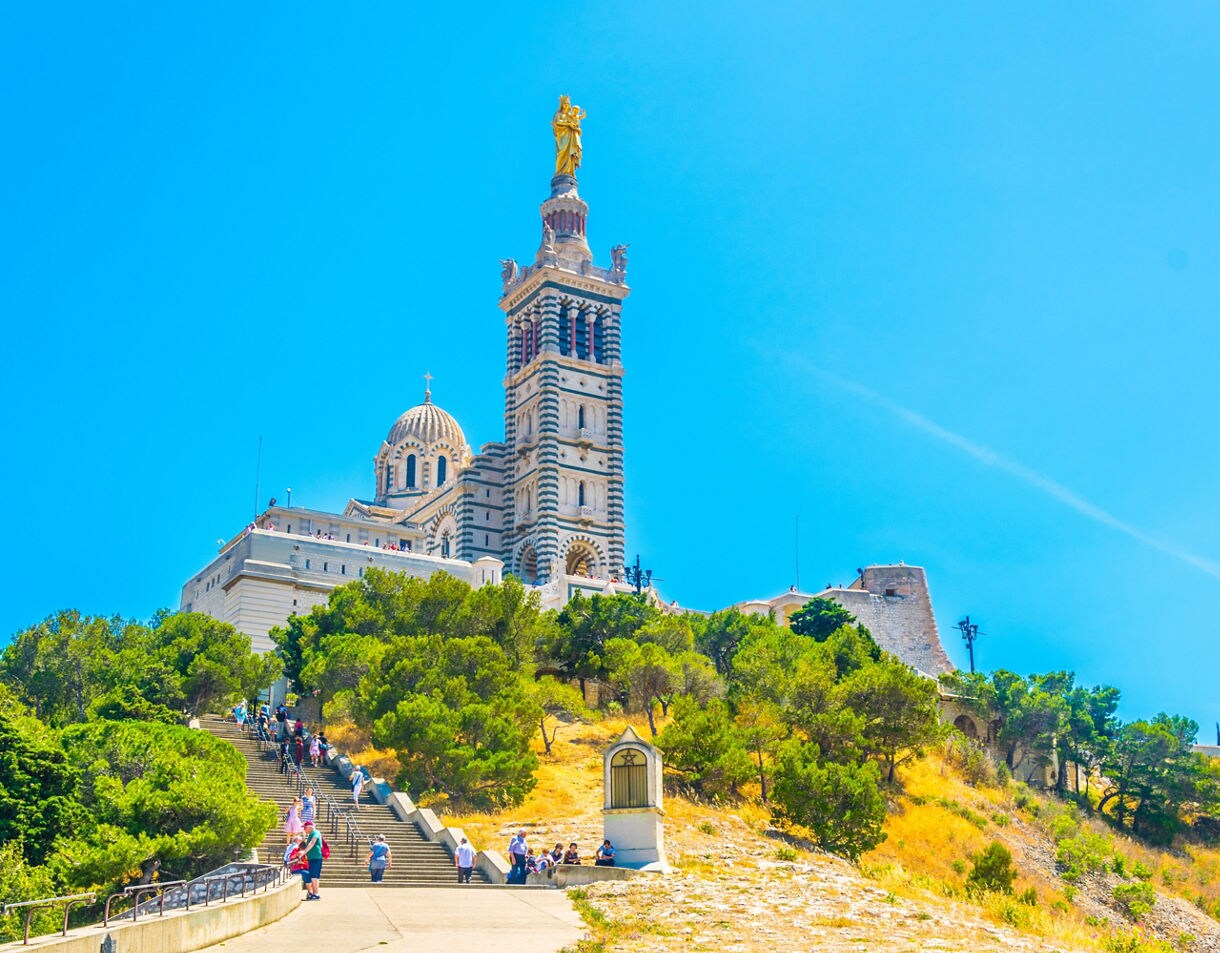 Notre-Dame de la Garde basilica rising above a sunlit hill with visitors walking up steps, blue sky overhead.