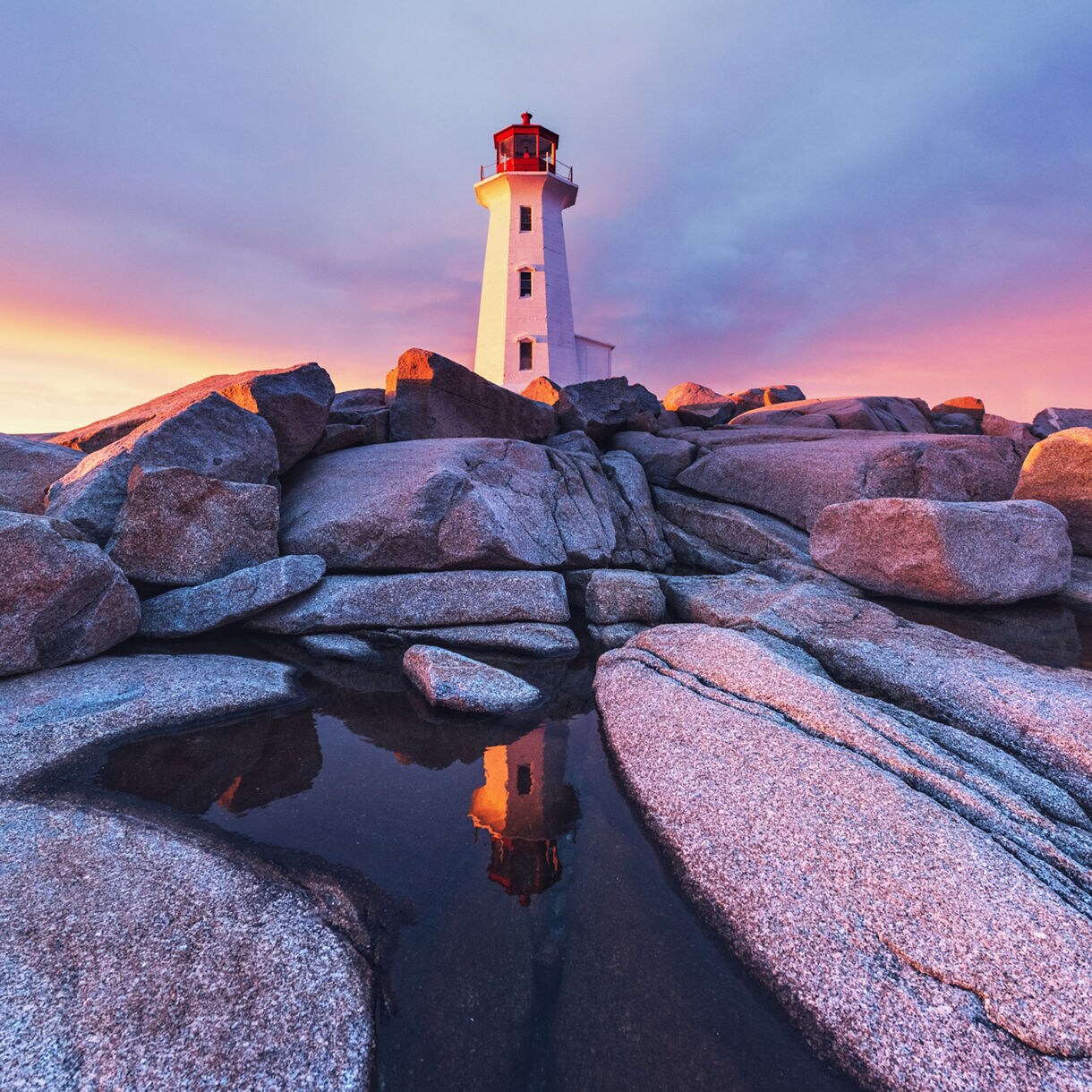 A lighthouse stands on rocky shores at sunset, with vibrant colors in the sky reflecting on calm waters