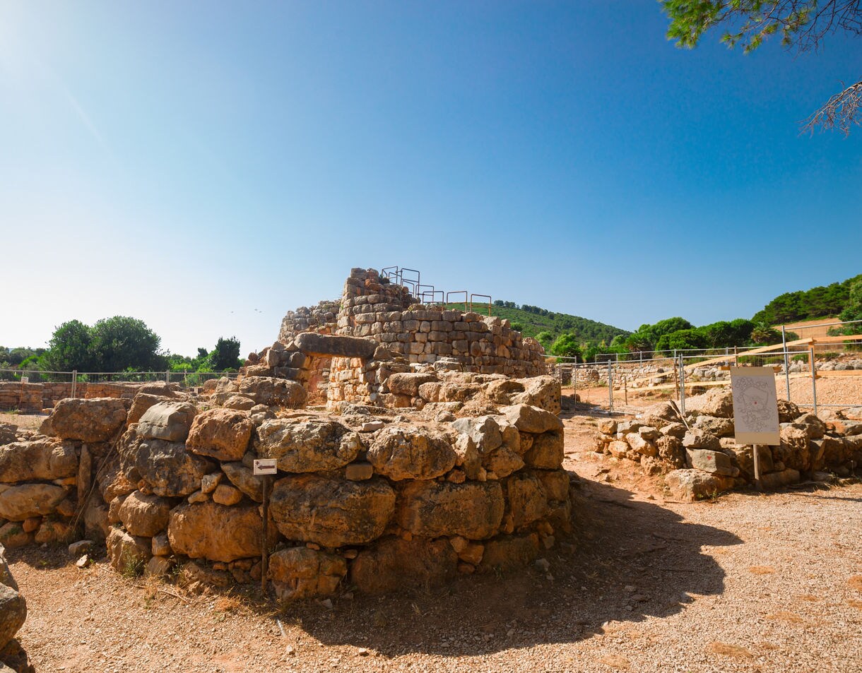 Stone ruins of a circular nuraghe complex in a dry landscape with hills and trees under a clear sky.