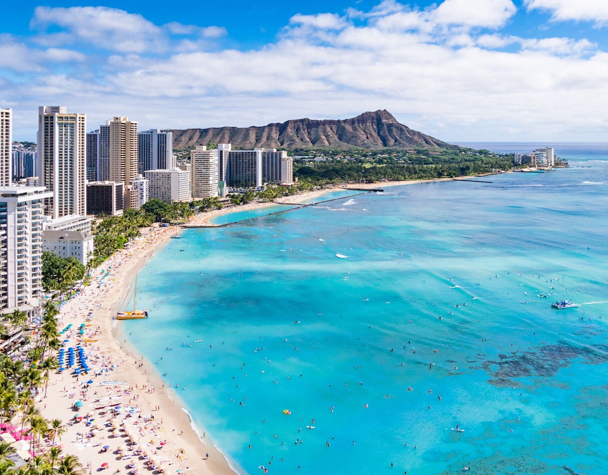 Aerial view of Waikiki Beach in Honolulu, lined with hotels and high-rises, with turquoise waters full of swimmers and surfers and Diamond Head crater in the distance.