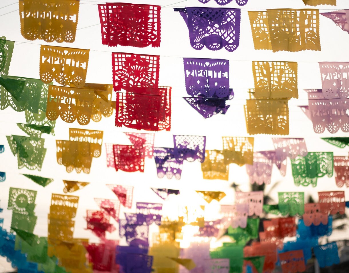 Colorful papel picado banners in red, yellow, green, purple and blue strung across the sky with sunlight shining through the cut-out designs.