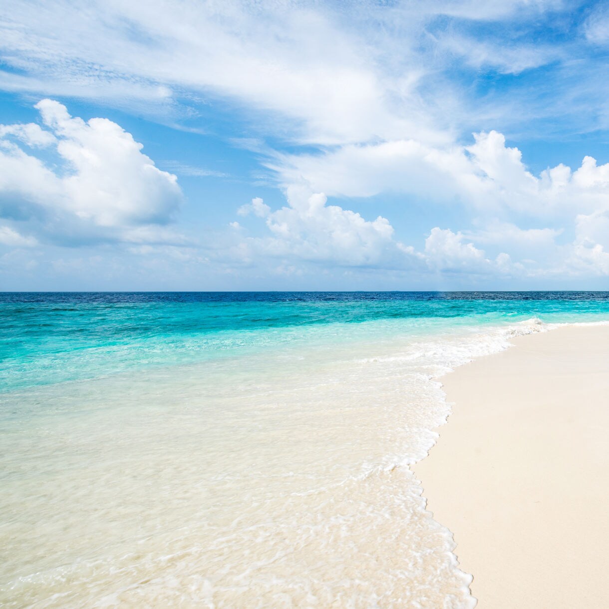 Peaceful view of a white sand beach in Okinawa with gentle waves, clear blue water and a bright sky filled with scattered clouds.