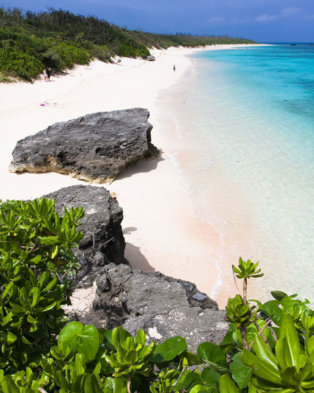 View of a quiet Okinawan beach with large dark rock formations, white sand, clear shallow water and dense green foliage under a partly cloudy sky.