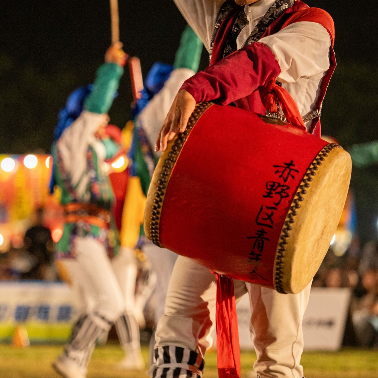 Close-up of a performer playing a traditional drum during an Okinawan Eisa dance, wearing colorful costume with blurred background of other dancers and festival lights.