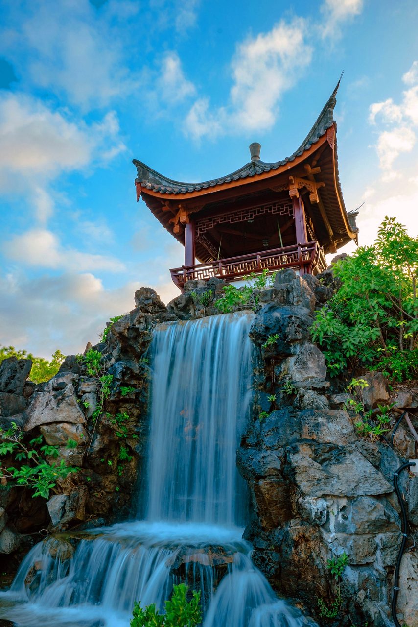 View of a red pagoda-style pavilion above a cascading waterfall surrounded by rocks and vibrant green plants in Okinawa’s Fukushen Garden.