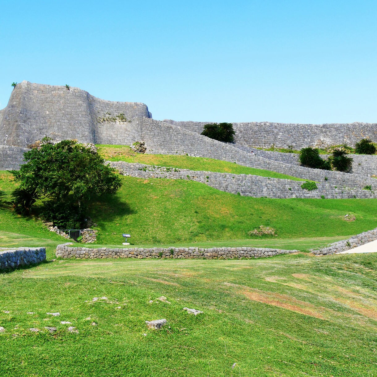 Wide view of Katsuren Castle ruins in Okinawa, featuring layered stone walls, green grassy slopes and a clear blue sky above.