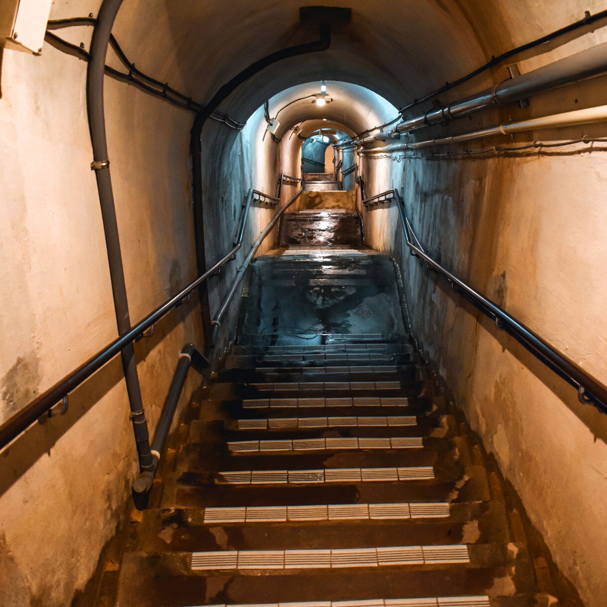 View down a dimly lit, narrow tunnel with wet stairs and exposed pipes inside the Former Japanese Navy Underground Headquarters in Okinawa.