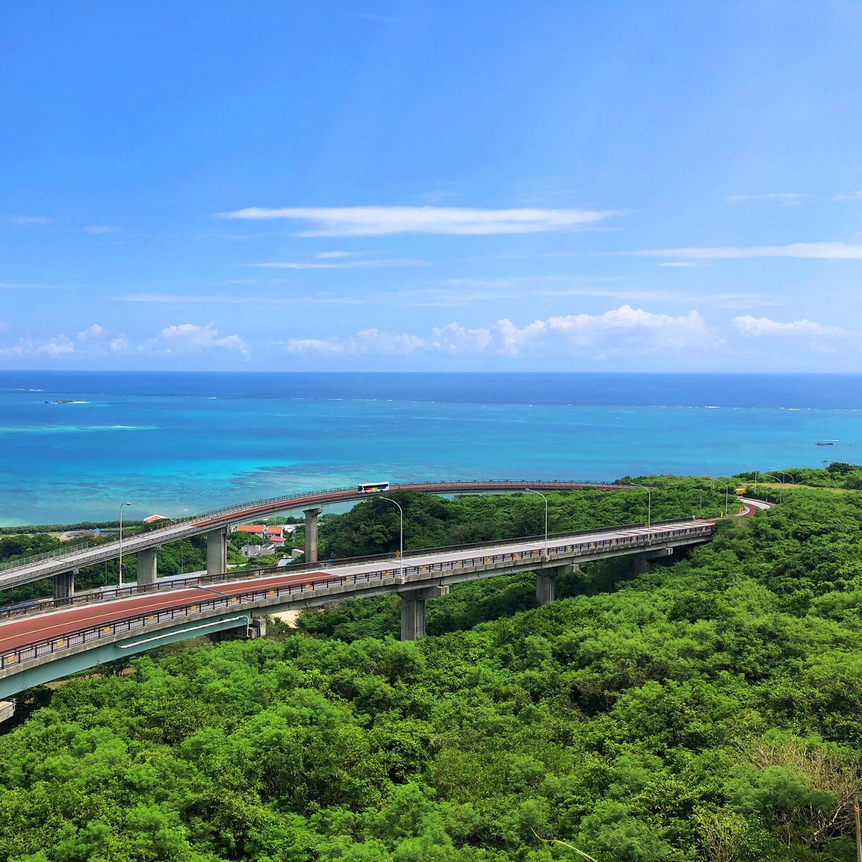 Elevated view of the winding Nirai Kanai Bridge in Okinawa, stretching over lush green hills with the clear blue sea and sky in the background