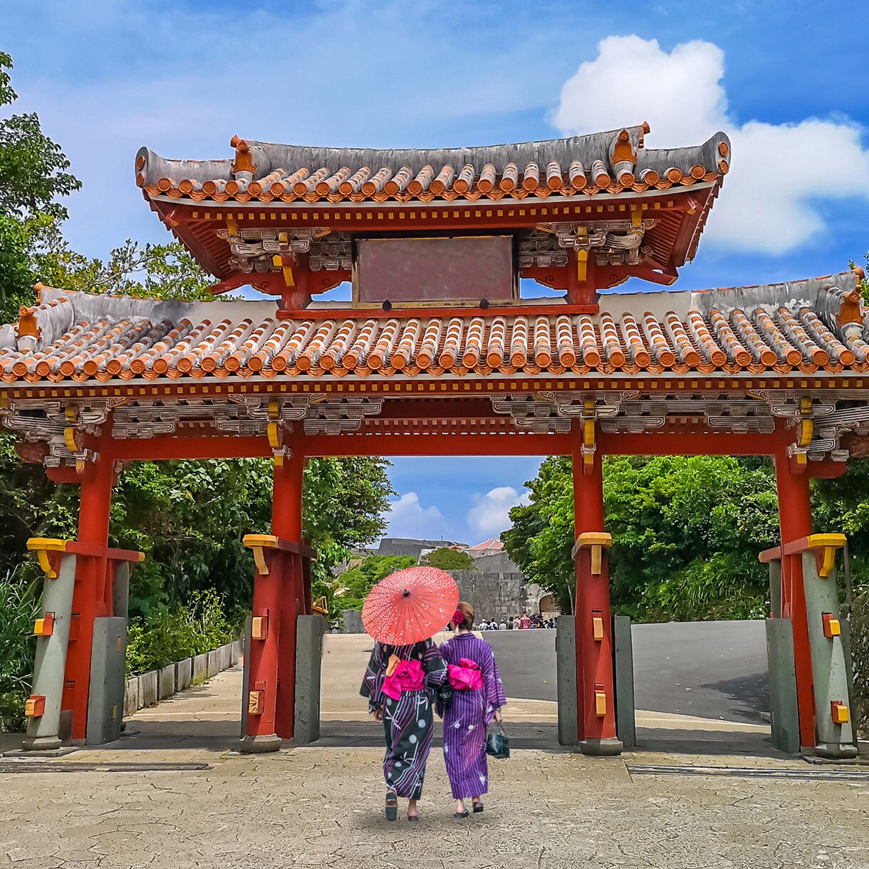 Two women in traditional kimonos walk under Okinawa’s Shureimon Gate holding a red parasol, surrounded by lush greenery and a bright blue sky.