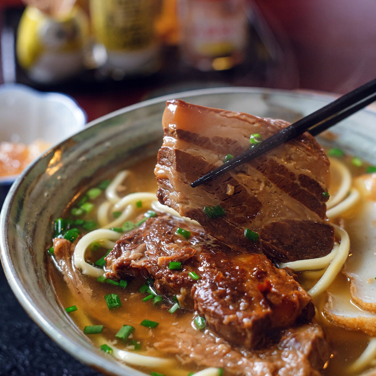 Close-up of a bowl of Okinawa soba with thick noodles, braised pork belly, sliced green onions and fish cake in hot broth, surrounded by side dishes including rice and pickled vegetables.