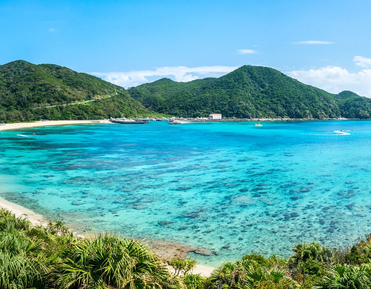 Panoramic view of a tropical Okinawan bay with lush green hills, clear blue water, coral reefs and white sand beaches dotted with visitors under a bright blue sky.