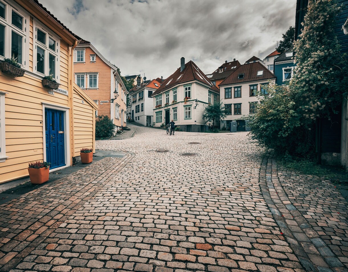 Cobblestone street in Bergen’s Old Town with pastel wooden houses and red-tiled roofs under cloudy skies.