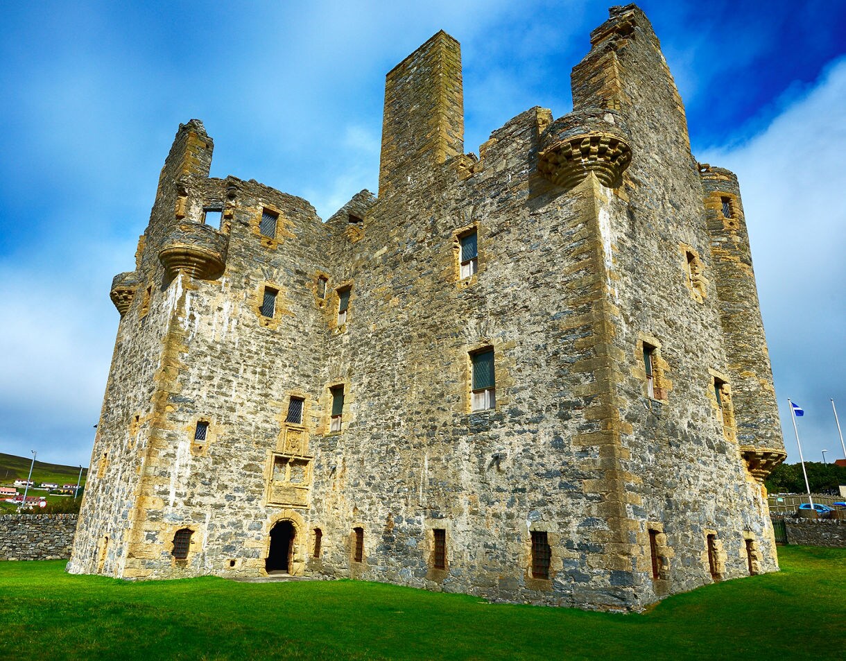 Stone walls and towers of Scalloway Castle in Shetland, standing tall against a bright blue sky with patches of clouds.
