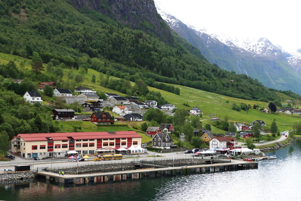 The port of Olden, Norway, with a dockside terminal, colorful houses and green hills rising to snow-dusted mountains in the background.