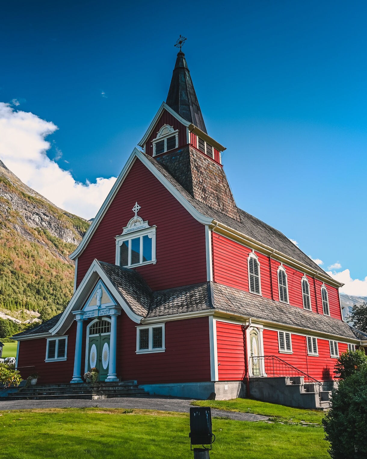 Bright red wooden church with a tall steeple in Olden, Norway, framed by green fields, trees and rugged mountains in the background.