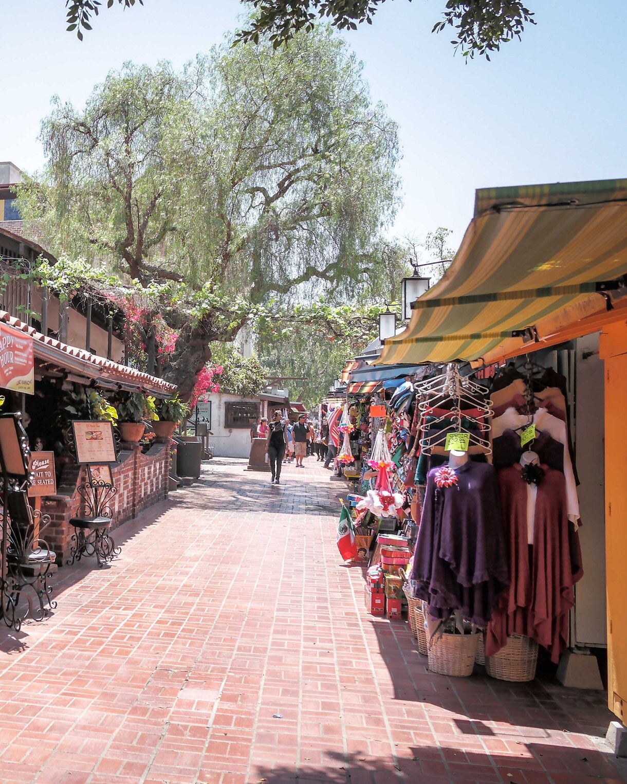 Brick walkway bordered by restaurant patios and stalls selling colorful goods under striped awnings.