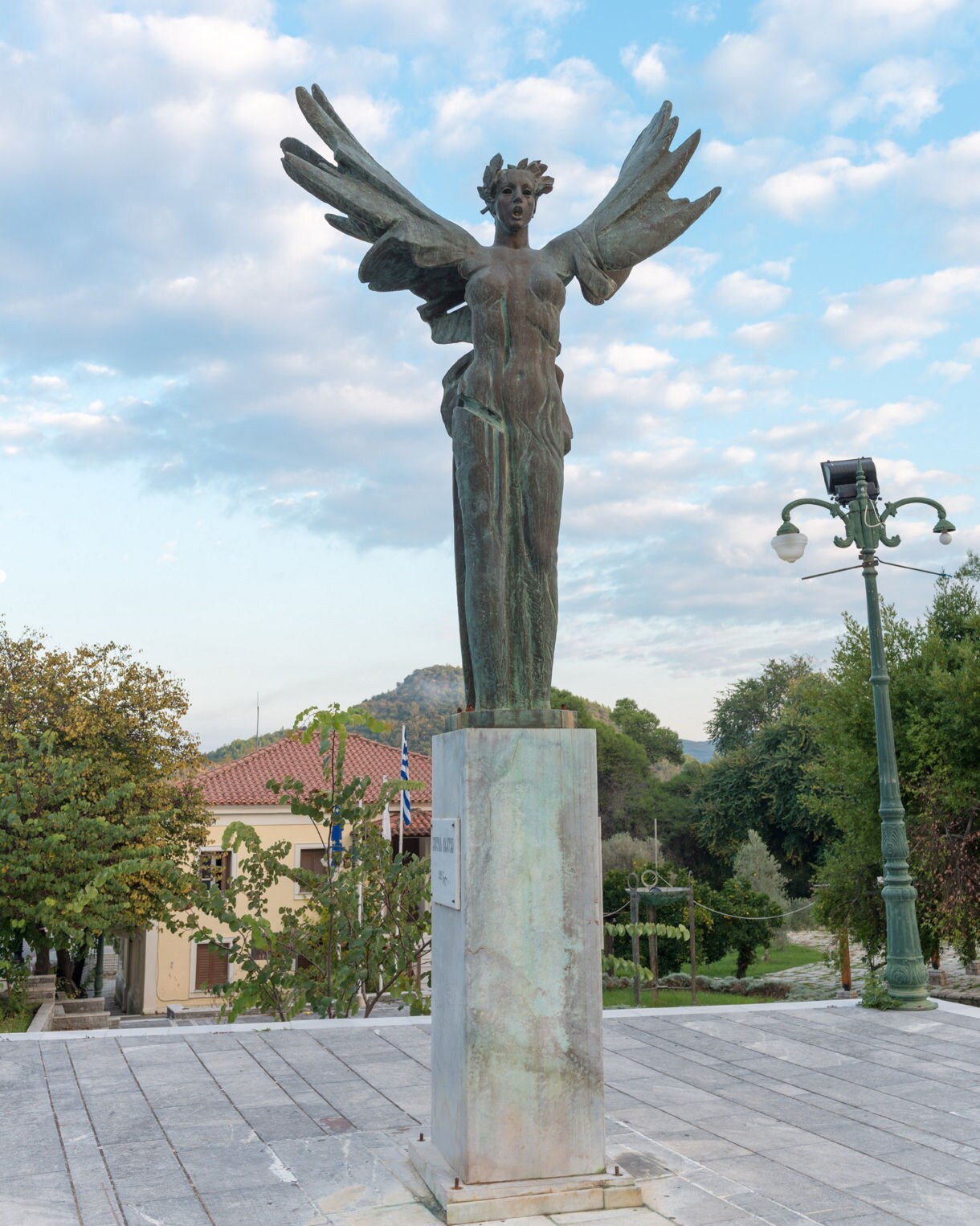 Bronze statue of a winged female figure in Olympia, Greece, standing on a stone pedestal with trees, rooftops and a cloudy blue sky in the background.