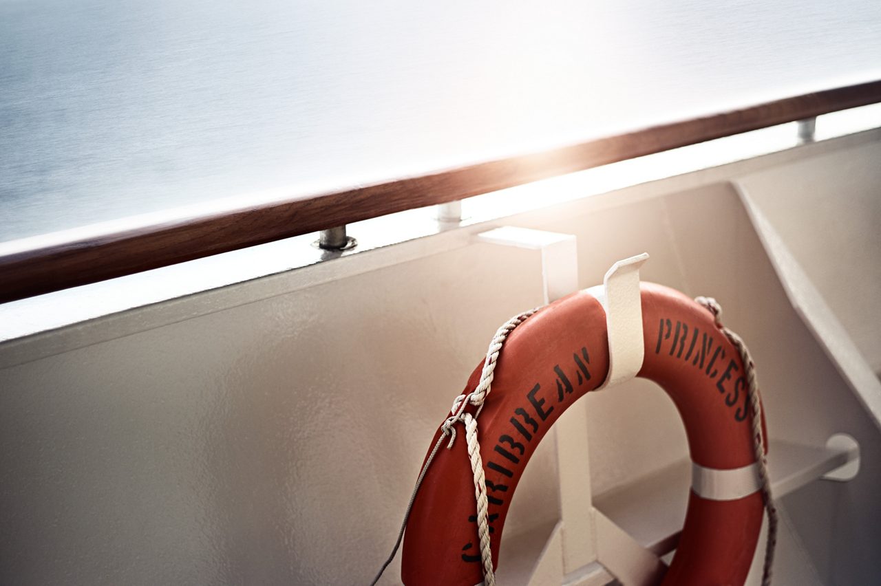 A red and white life ring labeled "Caribbean Princess" hangs on a cruise ship railing in bright sunlight.
