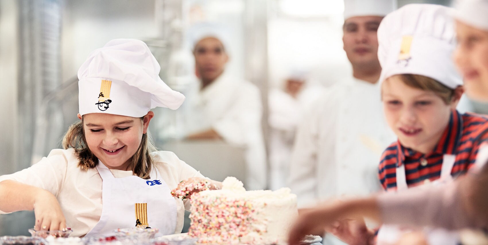 A group of children, wearing chef hats and aprons, are gathered around a table in a bright, professional kitchen. The main subject is a child actively decorating a large cake with colorful sprinkles. The atmosphere is lively and collaborative, with various baking ingredients and utensils visible. 