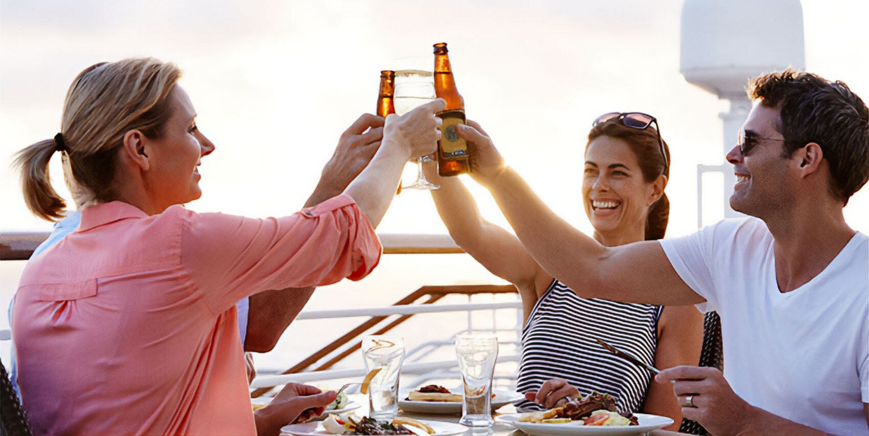 Three adults are raising beer bottles in a celebratory toast while seated at a table outdoors. The setting appears to be a deck or patio with a scenic view at sunset, featuring plates of food and glasses. The mood is relaxed and festive, with warm lighting and casual attire.