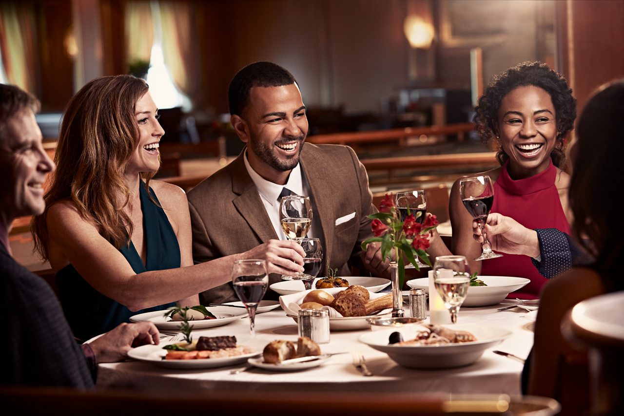 A couple dressed in elegant clothes siting at a table enjoying a meal and toasting glasses