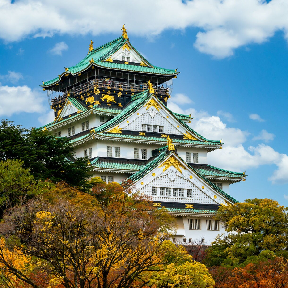 Osaka Castle with its green and gold roof stands tall above vibrant autumn foliage under a bright blue sky with scattered clouds.