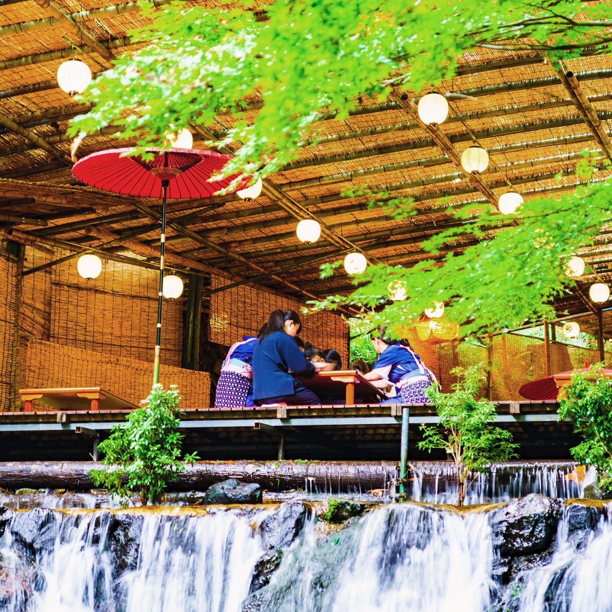 People in traditional attire enjoy a meal at a kawadoko dining platform above a small waterfall, surrounded by greenery and lit by lanterns under a bamboo roof.