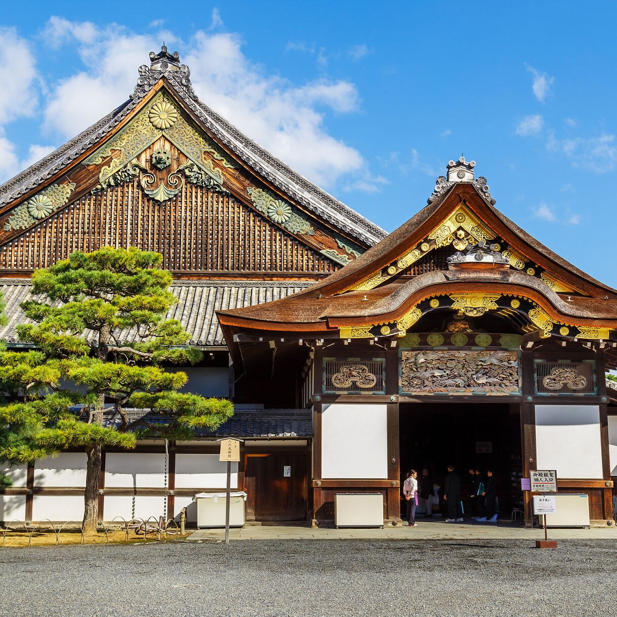Front view of Nijo Castle in Kyoto featuring detailed wooden architecture, golden embellishments and manicured pine trees under a bright blue sky.