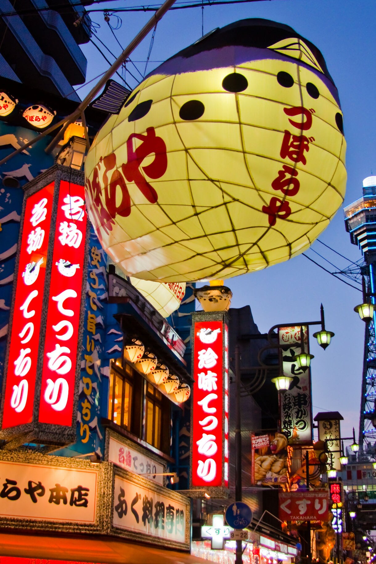 A large yellow paper lantern with red Japanese text and black polka dots hangs above a busy street lined with colorful neon signs and traditional lanterns, with Tsutenkaku Tower visible in the background against a purple twilight sky.