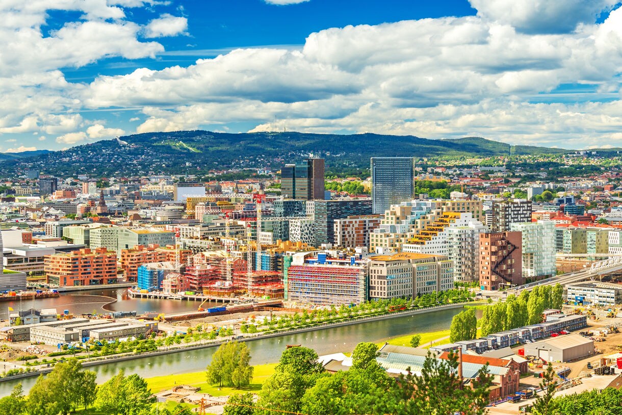 Aerial view of Oslo, Norway, showcasing modern buildings, construction cranes and tree-lined canals with green hills in the background under a partly cloudy sky.