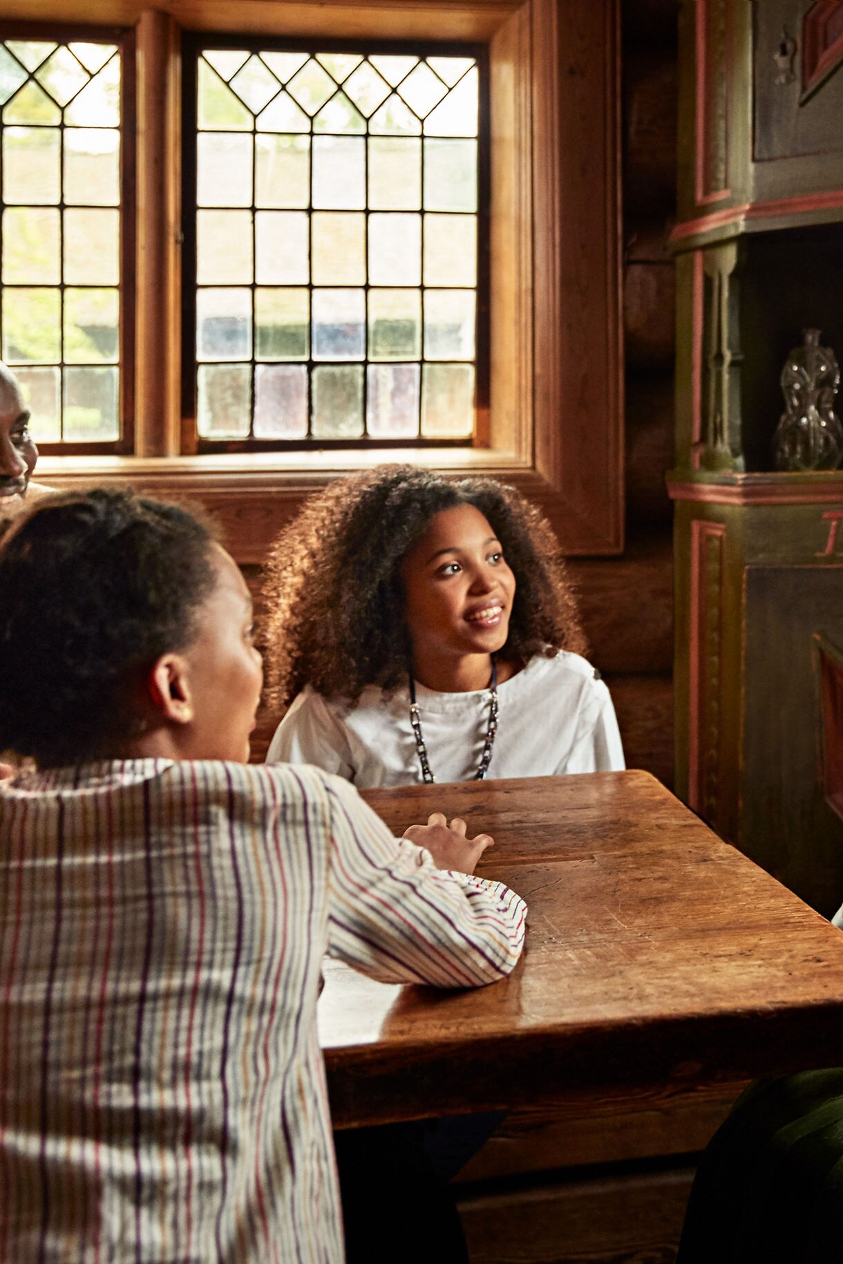 A family sits around a wooden table listening to a woman in traditional Norwegian folk costume inside a warm, rustic room with wooden walls and antique furniture.