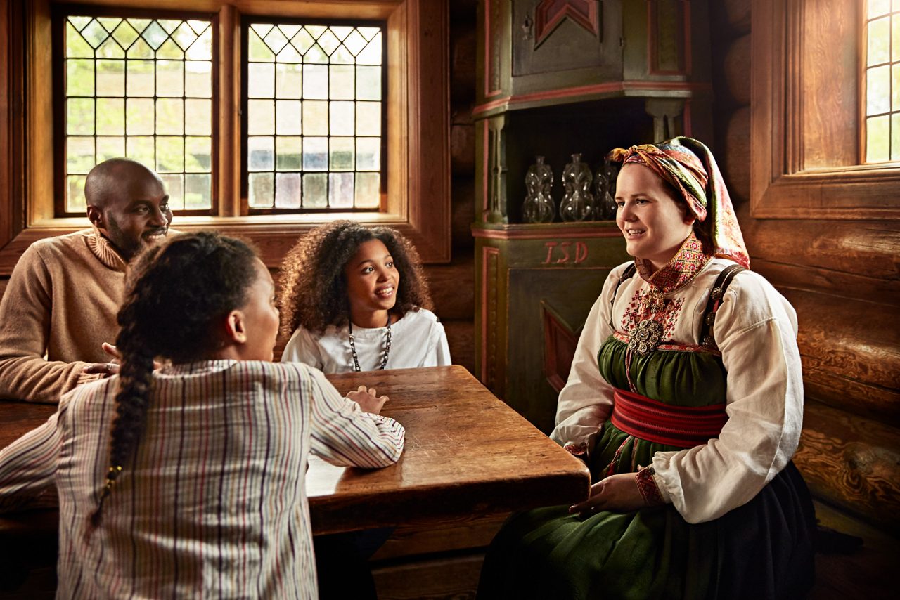 A family sits around a wooden table listening to a woman in traditional Norwegian folk costume inside a warm, rustic room with wooden walls and antique furniture.