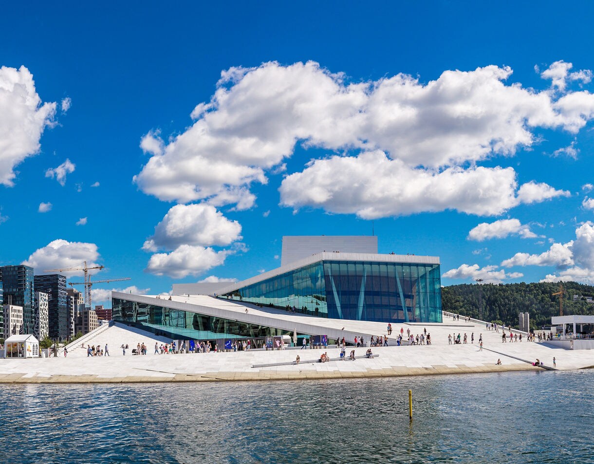 Oslo Opera House in Norway, with its angular white marble roof and glass facade overlooking the water under a bright blue sky with scattered clouds.