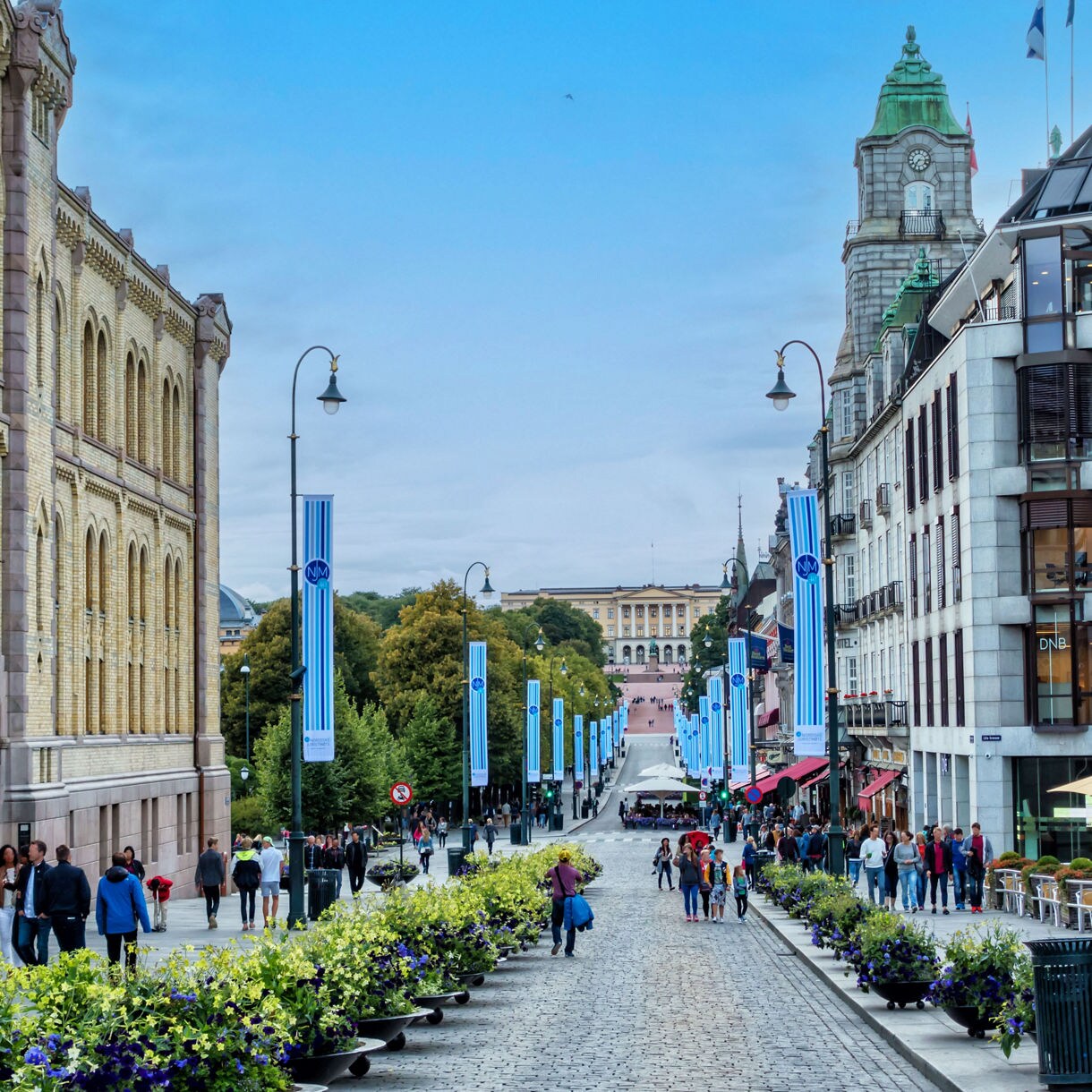 A bustling pedestrian street lined with flower planters, shops and historic buildings leads toward a distant palace in Oslo.