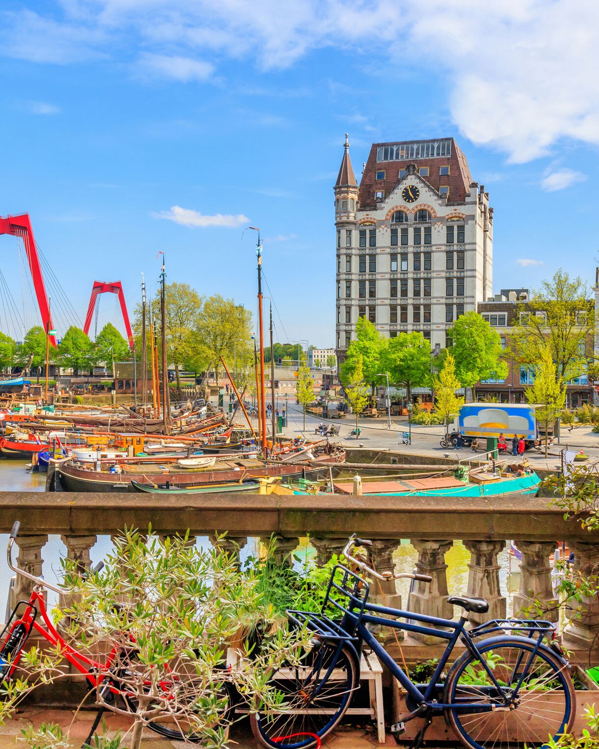 View of Rotterdam’s Oude Haven with moored boats, the red Willemsbrug bridge in the background and the White House building framed by spring trees and bright blue sky.