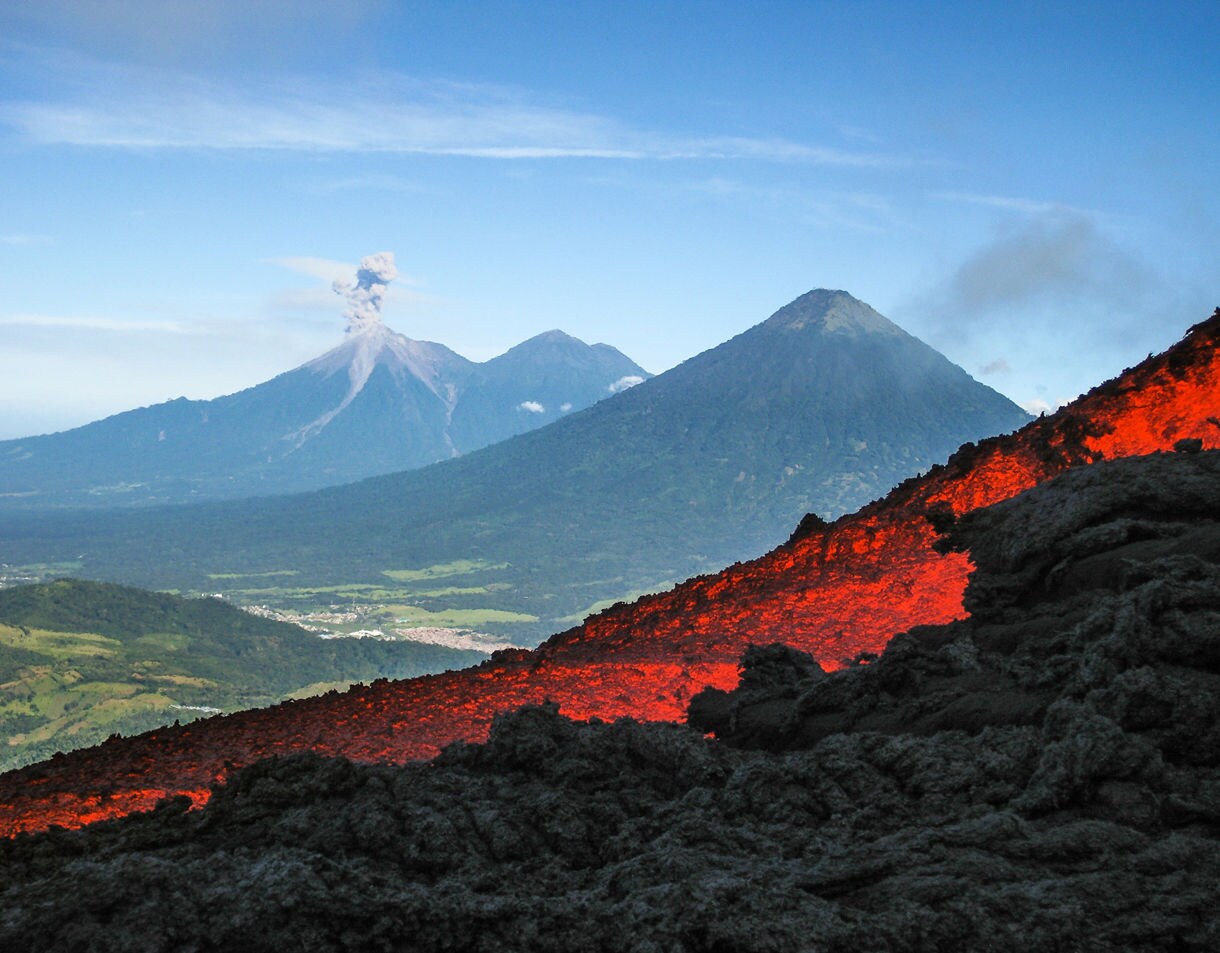 Flowing lava on the slopes of Pacaya Volcano in Guatemala with two nearby volcanoes visible in the distance, one releasing a plume of ash under a clear blue sky.