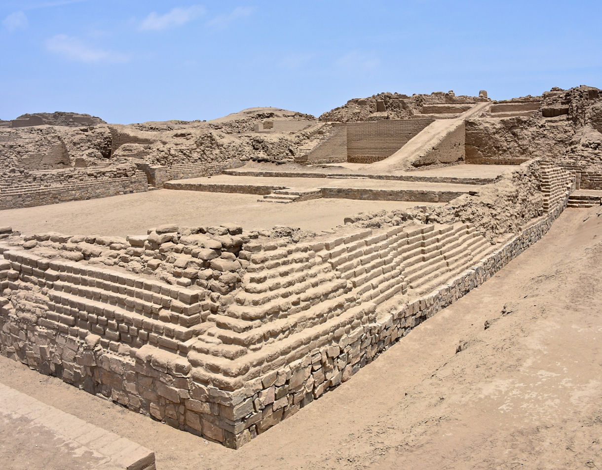 Wide view of the Pachacamac ruins, showing layered adobe platforms, stone foundations and sandy pathways under a bright blue sky.