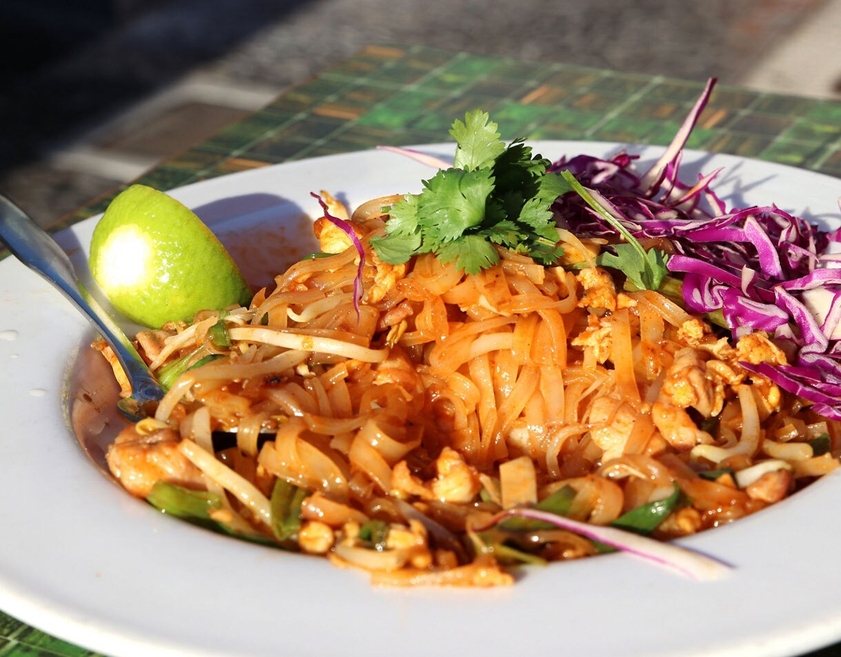 Close-up of a plate of pad Thai topped with cilantro, shredded purple cabbage and a lime wedge, with noodles mixed with vegetables and pieces of chicken.