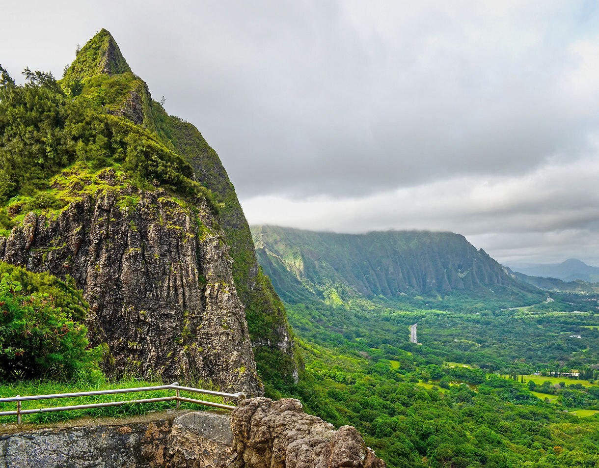 View from Pali Lookout on Oahu showing steep green cliffs, mist-covered ridges and a wide valley below with winding roads and scattered towns.
