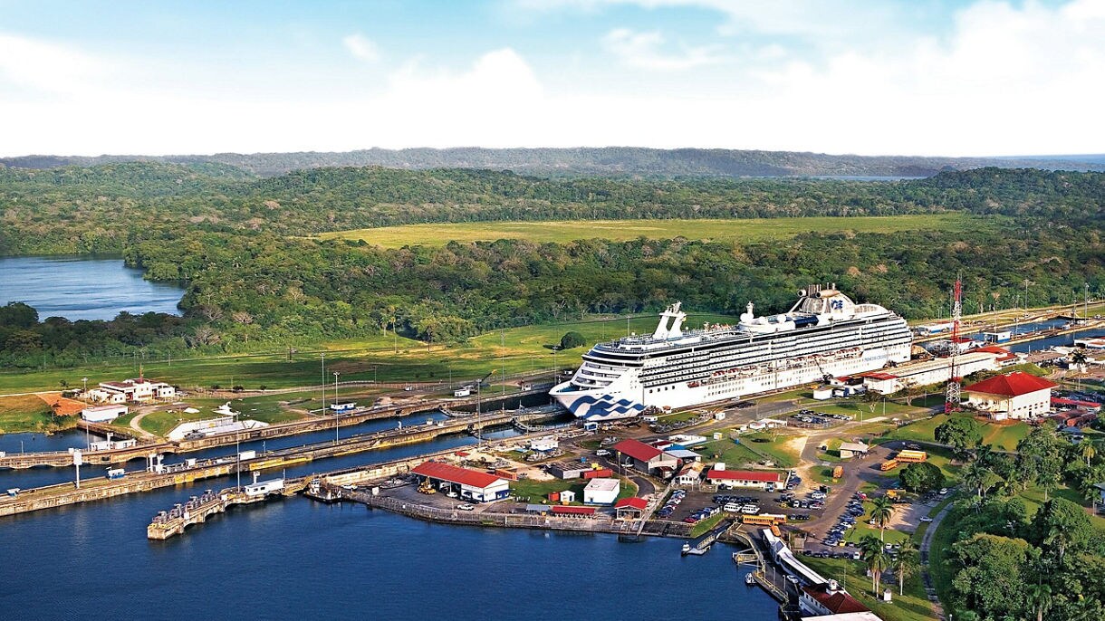 An aerial view of a Princess cruise ship passing through the Panama Canal locks, surrounded by green jungle hills and canal infrastructure.